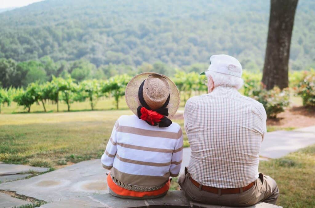 An older man and woman sit on a picnic blanket and look out over a rolling green landscape.