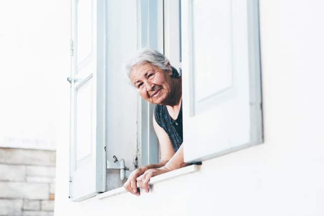 A senior-aged woman peers out her balcony window, smiling. Photo by Nick Karvounis on Unsplash.