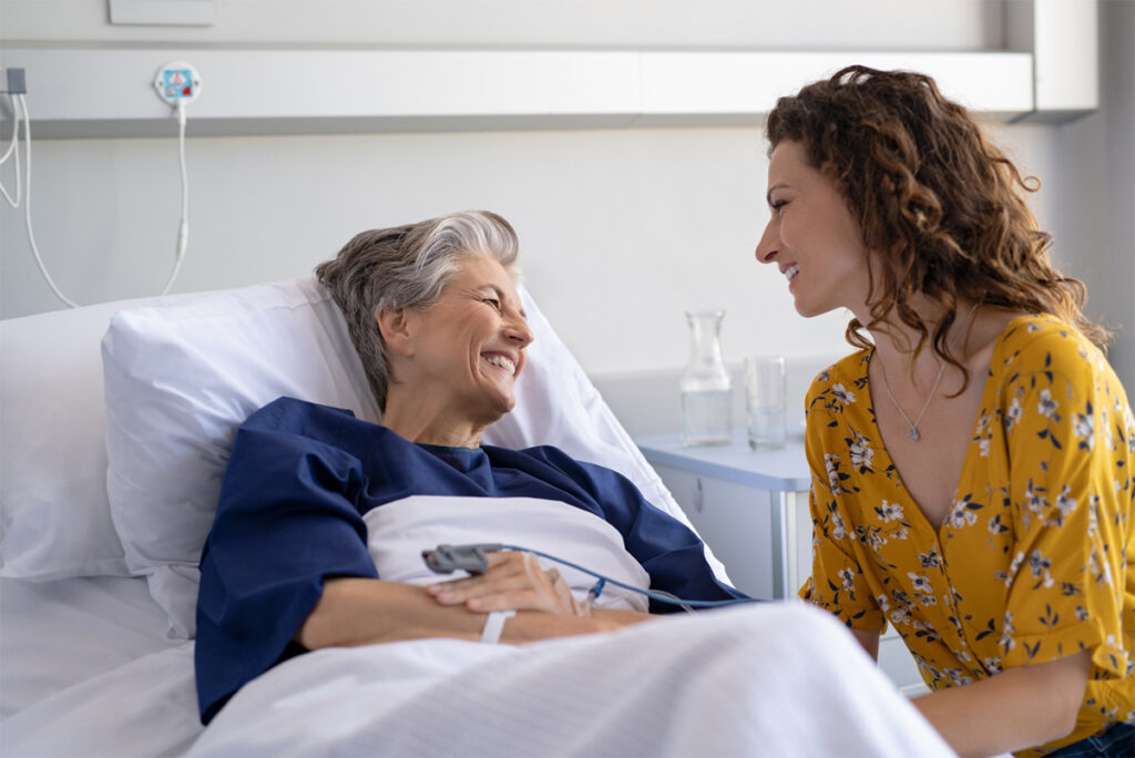 A senior woman lying in a hospital bed while speaking with a family member sitting next to the bed
